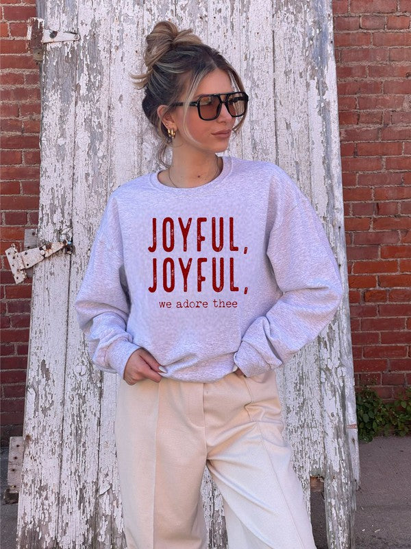 A person wearing a beige beanie and a Joyful, Joyful, We Adore Thee Sweatshirt made of 50% cotton and pre-shrunk fleece sits with a smiling expression, making a peace sign with both hands.