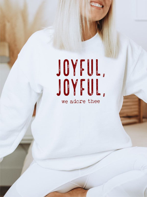 A person wearing a beige beanie and a Joyful, Joyful, We Adore Thee Sweatshirt made of 50% cotton and pre-shrunk fleece sits with a smiling expression, making a peace sign with both hands.