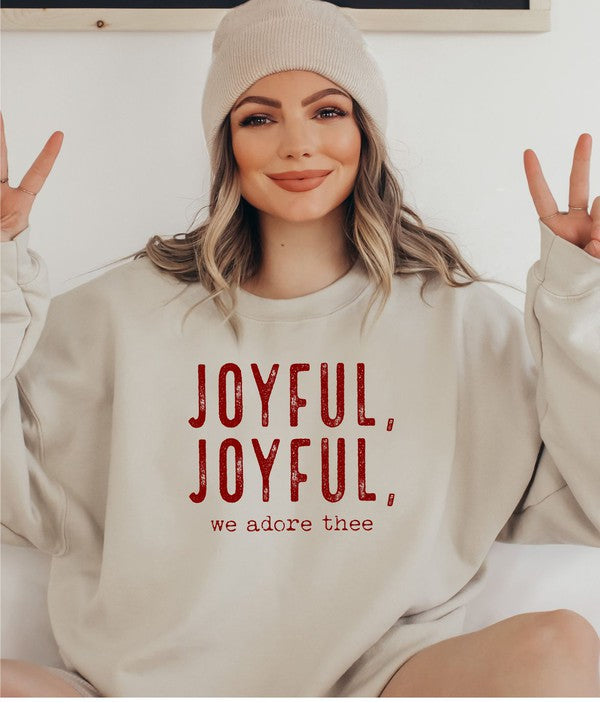 A person wearing a beige beanie and a Joyful, Joyful, We Adore Thee Sweatshirt made of 50% cotton and pre-shrunk fleece sits with a smiling expression, making a peace sign with both hands.