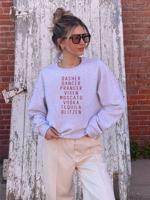 A person stands in front of a weathered wooden door, wearing the Dasher Dancer Prancer Vixen Boutique Sweatshirt, which is a light gray crew neck with maroon print listing reindeer and alcoholic beverages. They are also dressed in light-colored pants and large black sunglasses.