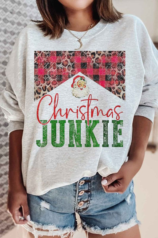 A person is seen wearing denim shorts paired with a CHRISTMAS JUNKIE GRAPHIC SWEATSHIRT, which boasts festive graphics such as a Santa face, leopard print, and a plaid pattern. Crafted from premium cotton and designed with unisex sizing to ensure the perfect fit.