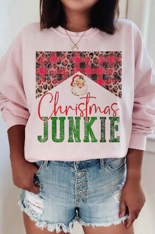 A person is seen wearing denim shorts paired with a CHRISTMAS JUNKIE GRAPHIC SWEATSHIRT, which boasts festive graphics such as a Santa face, leopard print, and a plaid pattern. Crafted from premium cotton and designed with unisex sizing to ensure the perfect fit.