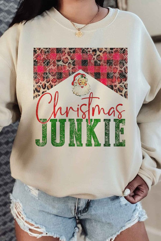 A person is seen wearing denim shorts paired with a CHRISTMAS JUNKIE GRAPHIC SWEATSHIRT, which boasts festive graphics such as a Santa face, leopard print, and a plaid pattern. Crafted from premium cotton and designed with unisex sizing to ensure the perfect fit.