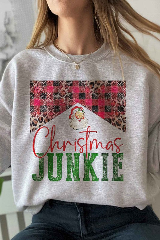 A person is seen wearing denim shorts paired with a CHRISTMAS JUNKIE GRAPHIC SWEATSHIRT, which boasts festive graphics such as a Santa face, leopard print, and a plaid pattern. Crafted from premium cotton and designed with unisex sizing to ensure the perfect fit.