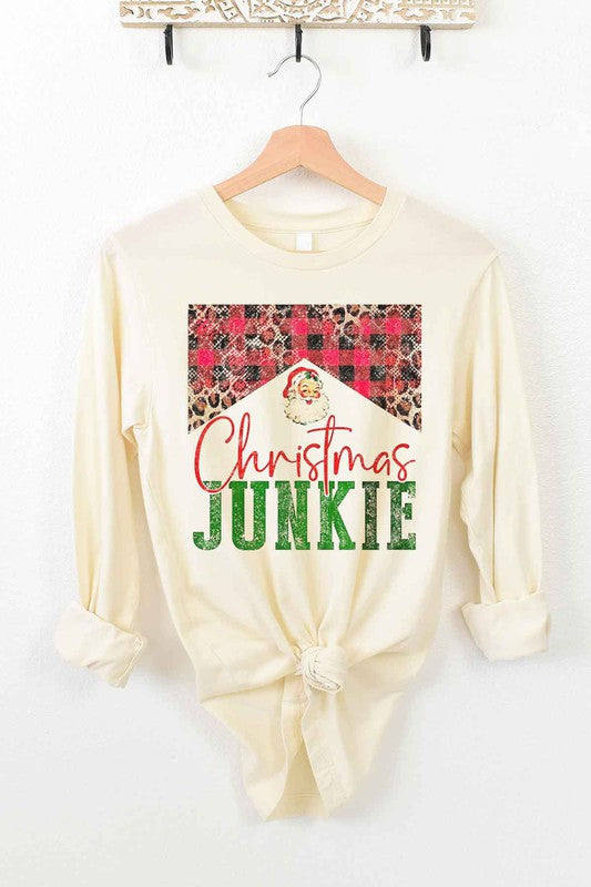 The CHRISTMAS JUNKIE LONG SLEEVE TEE, made from premium cotton, hangs gracefully on a wooden hanger. It features a beige color and showcases a festive design with the words "Christmas Junkie" in vibrant red and green text. The tee is adorned with cheerful plaid and leopard print patterns, making it perfect for celebrating the holiday spirit.