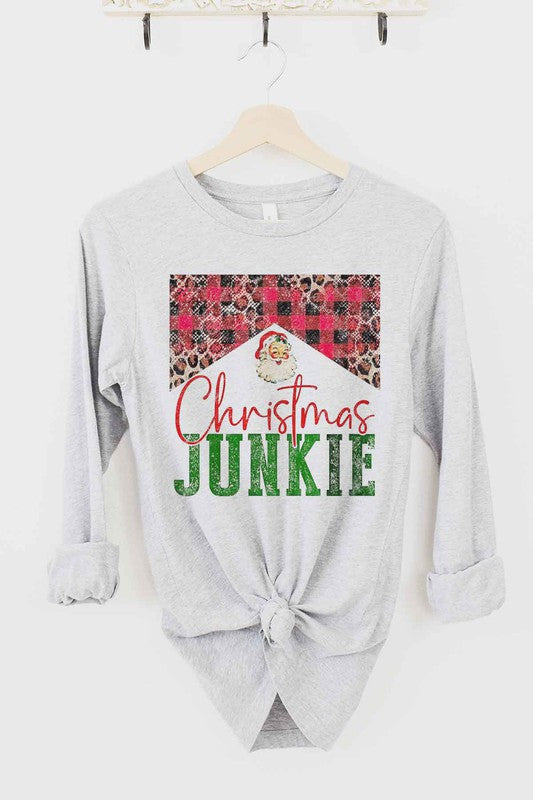 The CHRISTMAS JUNKIE LONG SLEEVE TEE, made from premium cotton, hangs gracefully on a wooden hanger. It features a beige color and showcases a festive design with the words "Christmas Junkie" in vibrant red and green text. The tee is adorned with cheerful plaid and leopard print patterns, making it perfect for celebrating the holiday spirit.