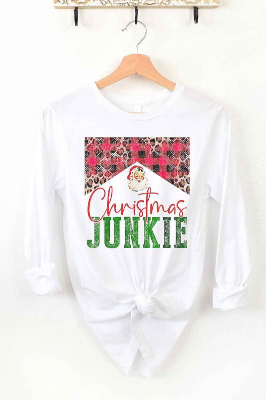 The CHRISTMAS JUNKIE LONG SLEEVE TEE, made from premium cotton, hangs gracefully on a wooden hanger. It features a beige color and showcases a festive design with the words "Christmas Junkie" in vibrant red and green text. The tee is adorned with cheerful plaid and leopard print patterns, making it perfect for celebrating the holiday spirit.