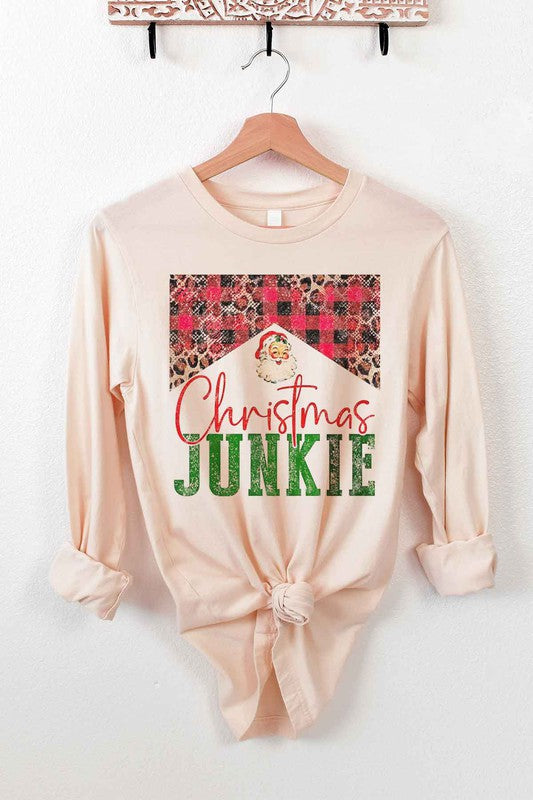 The CHRISTMAS JUNKIE LONG SLEEVE TEE, made from premium cotton, hangs gracefully on a wooden hanger. It features a beige color and showcases a festive design with the words "Christmas Junkie" in vibrant red and green text. The tee is adorned with cheerful plaid and leopard print patterns, making it perfect for celebrating the holiday spirit.