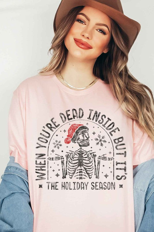 A young woman with long hair in a pink hat is wearing the CHRISTMAS SKELETON GRAPHIC TEE / T-SHIRT, crafted from premium cotton, featuring a white base with the text "When you're dead inside but it's the holiday season.