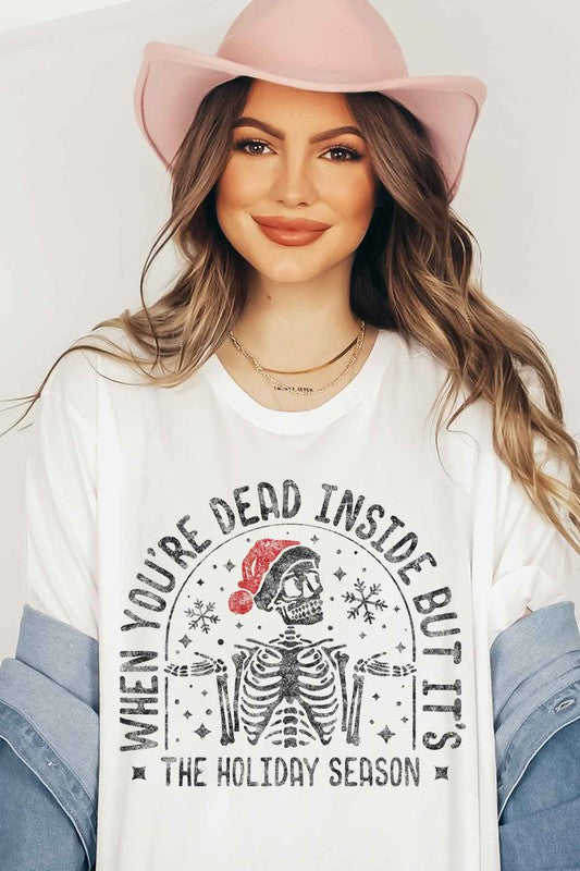 A young woman with long hair in a pink hat is wearing the CHRISTMAS SKELETON GRAPHIC TEE / T-SHIRT, crafted from premium cotton, featuring a white base with the text "When you're dead inside but it's the holiday season.