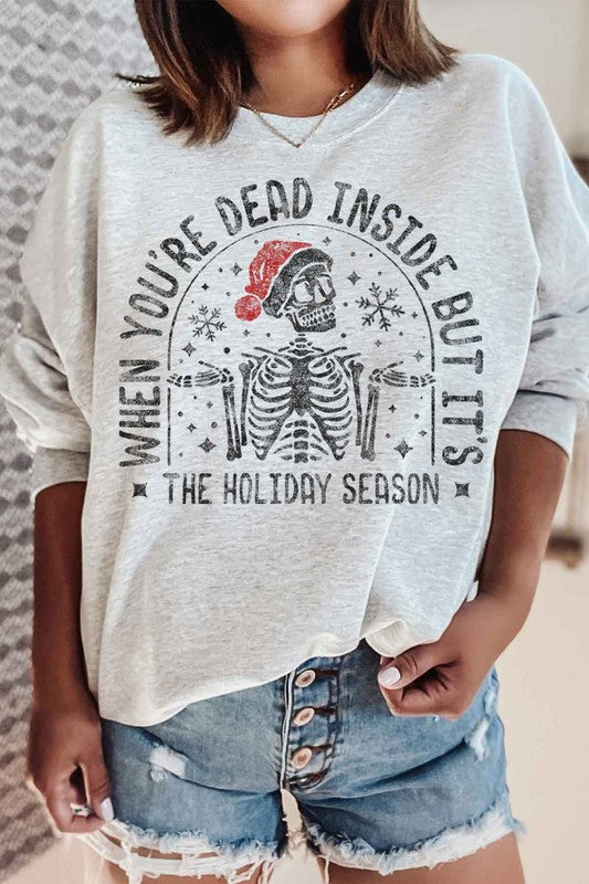 Person wearing the Christmas Skeleton Graphic Sweatshirt in premium cotton gray, which features a festive skeleton with a Santa hat and the text "When you’re dead inside, but it’s the holiday season." A perfect addition to your holiday wardrobe.