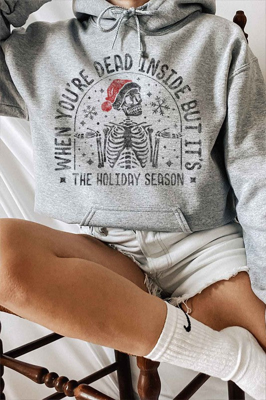 A person sits on a chair wearing the CHRISTMAS SKELETON GRAPHIC HOODIE, a premium cotton white hoodie with a Christmas skeleton graphic and Santa hat. The caption reads, "When you're dead inside but it's the holiday season." They have paired it with light shorts and white socks. Unisex sizing ensures everyone can enjoy the festive spirit.