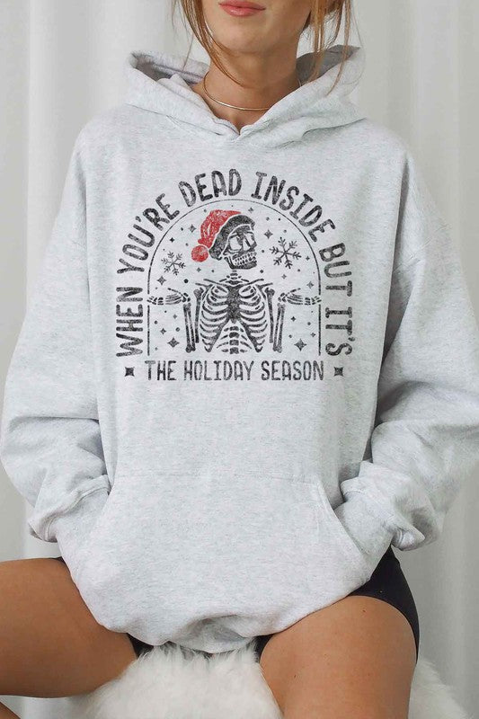 A person sits on a chair wearing the CHRISTMAS SKELETON GRAPHIC HOODIE, a premium cotton white hoodie with a Christmas skeleton graphic and Santa hat. The caption reads, "When you're dead inside but it's the holiday season." They have paired it with light shorts and white socks. Unisex sizing ensures everyone can enjoy the festive spirit.