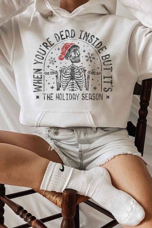 A person sits on a chair wearing the CHRISTMAS SKELETON GRAPHIC HOODIE, a premium cotton white hoodie with a Christmas skeleton graphic and Santa hat. The caption reads, "When you're dead inside but it's the holiday season." They have paired it with light shorts and white socks. Unisex sizing ensures everyone can enjoy the festive spirit.