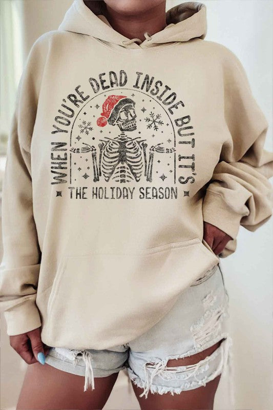 A person sits on a chair wearing the CHRISTMAS SKELETON GRAPHIC HOODIE, a premium cotton white hoodie with a Christmas skeleton graphic and Santa hat. The caption reads, "When you're dead inside but it's the holiday season." They have paired it with light shorts and white socks. Unisex sizing ensures everyone can enjoy the festive spirit.