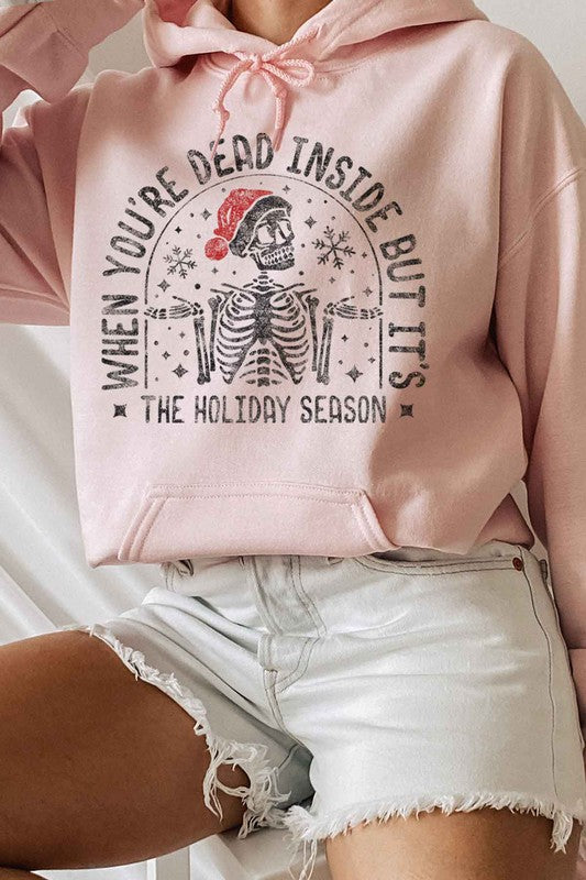 A person wearing a CHRISTMAS SKELETON HOODIE PLUS SIZE, featuring a festive skeleton graphic and the text "When you're dead inside but it's the holiday season," is seated on a chair in white shorts and socks, enjoying the comfort of premium cotton.