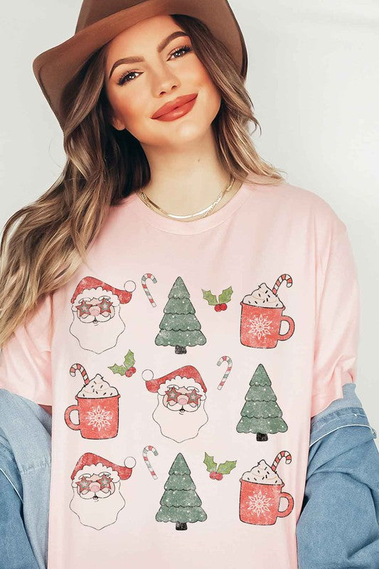 A woman wearing a pink hat and a white CHRISTMAS COLLECTION GRAPHIC TEE / T-SHIRT with festive holiday prints of Santa Claus, Christmas trees, candy canes, and mugs of hot chocolate stands against a light background. This classic fit is part of our exclusive Christmas collection.