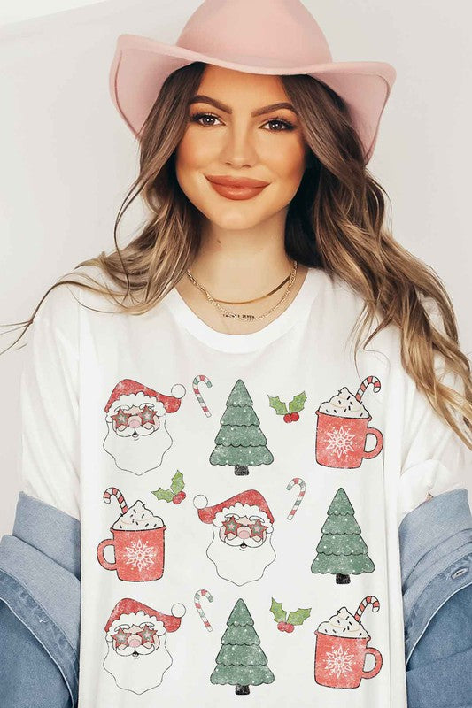 A woman wearing a pink hat and a white CHRISTMAS COLLECTION GRAPHIC TEE / T-SHIRT with festive holiday prints of Santa Claus, Christmas trees, candy canes, and mugs of hot chocolate stands against a light background. This classic fit is part of our exclusive Christmas collection.