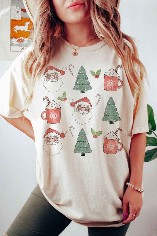 A woman wearing a pink hat and a white CHRISTMAS COLLECTION GRAPHIC TEE / T-SHIRT with festive holiday prints of Santa Claus, Christmas trees, candy canes, and mugs of hot chocolate stands against a light background. This classic fit is part of our exclusive Christmas collection.