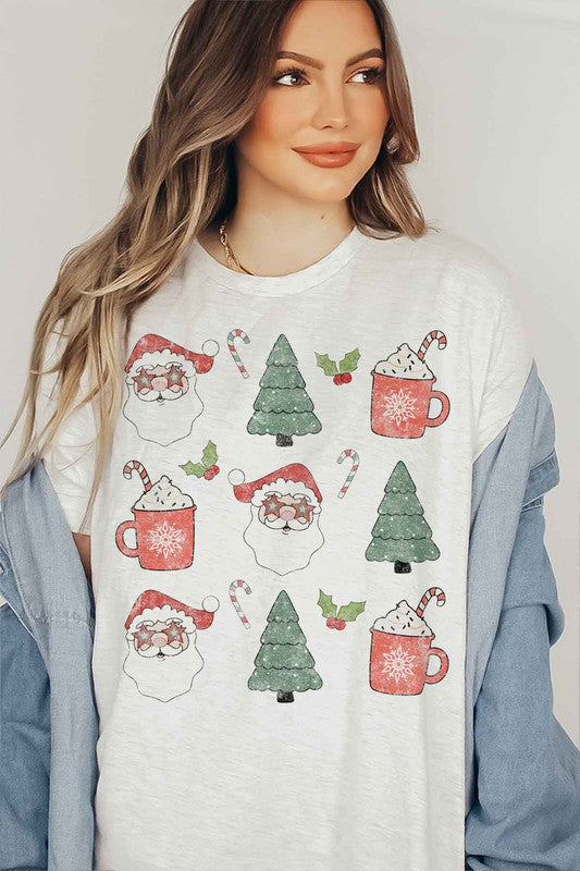 A woman wearing a pink hat and a white CHRISTMAS COLLECTION GRAPHIC TEE / T-SHIRT with festive holiday prints of Santa Claus, Christmas trees, candy canes, and mugs of hot chocolate stands against a light background. This classic fit is part of our exclusive Christmas collection.