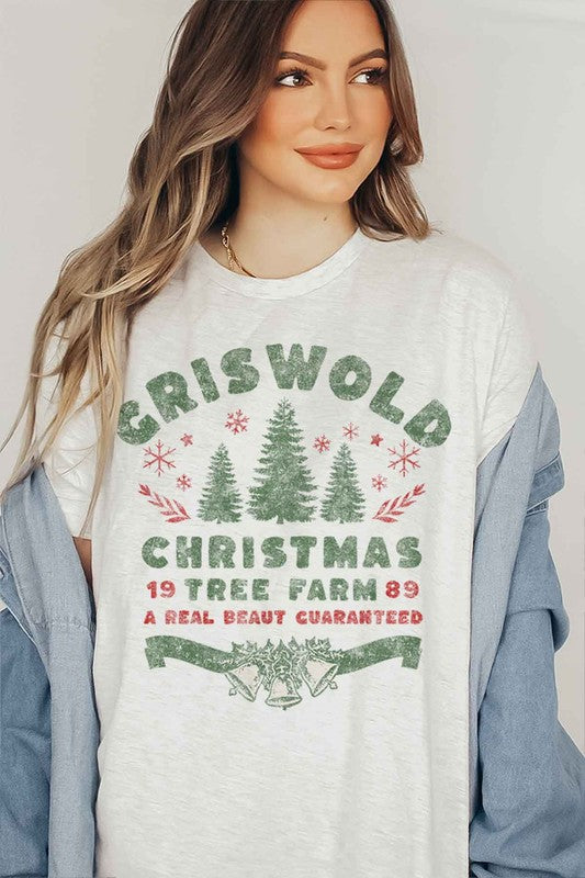 A woman wearing a pink hat and a CHRISTMAS TREE FARM GRAPHIC TEE that reads, "Criswold Christmas Tree Farm '89 - A Real Beaut Guaranteed," featuring Christmas trees and festive designs.