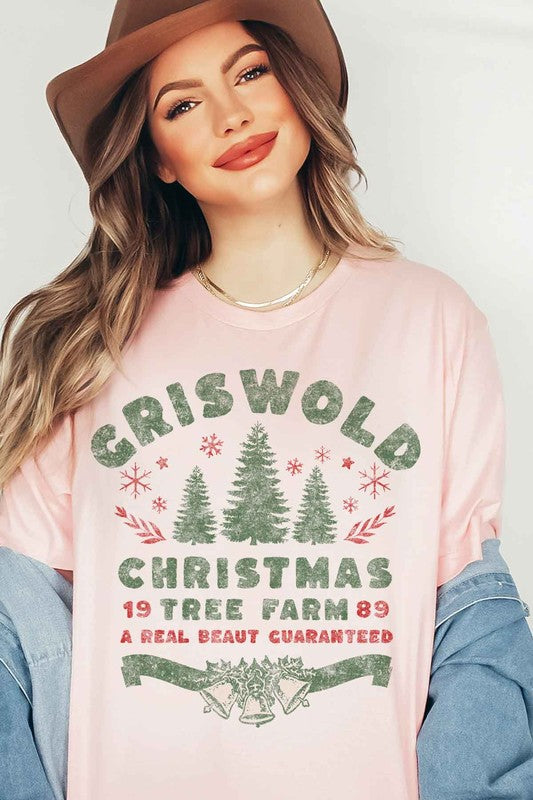 A woman wearing a pink hat and a CHRISTMAS TREE FARM GRAPHIC TEE that reads, "Criswold Christmas Tree Farm '89 - A Real Beaut Guaranteed," featuring Christmas trees and festive designs.