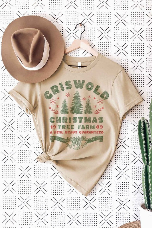 A woman wearing a pink hat and a CHRISTMAS TREE FARM GRAPHIC TEE that reads, "Criswold Christmas Tree Farm '89 - A Real Beaut Guaranteed," featuring Christmas trees and festive designs.