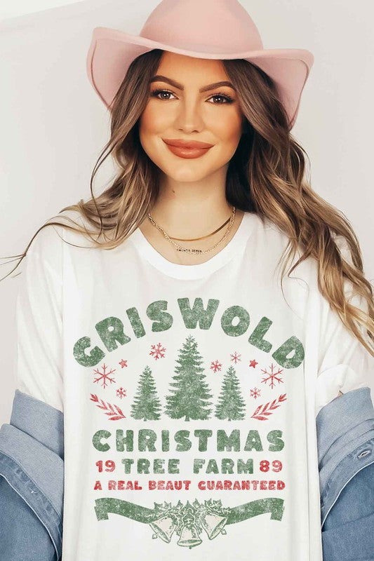 A woman wearing a pink hat and a CHRISTMAS TREE FARM GRAPHIC TEE that reads, "Criswold Christmas Tree Farm '89 - A Real Beaut Guaranteed," featuring Christmas trees and festive designs.