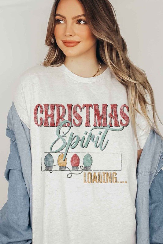 A person wearing a LOADING CHRISTMAS SPIRIT GRAPHIC TEE / T-SHIRT in beige, featuring the text "Christmas Spirit Loading..." and a festive holiday lights graphic, standing casually with one hand on their hip.