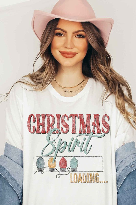 A person wearing a LOADING CHRISTMAS SPIRIT GRAPHIC TEE / T-SHIRT in beige, featuring the text "Christmas Spirit Loading..." and a festive holiday lights graphic, standing casually with one hand on their hip.