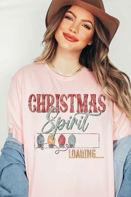 A person wearing a LOADING CHRISTMAS SPIRIT GRAPHIC TEE / T-SHIRT in beige, featuring the text "Christmas Spirit Loading..." and a festive holiday lights graphic, standing casually with one hand on their hip.