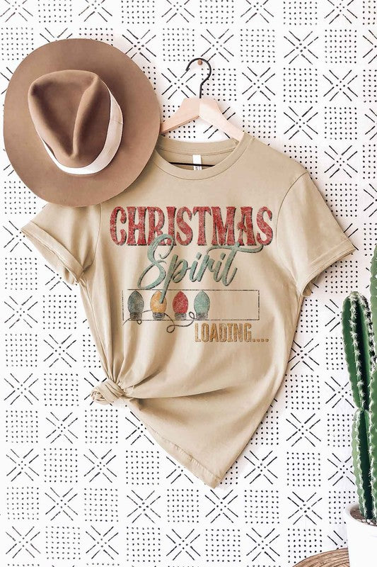 A person wearing a LOADING CHRISTMAS SPIRIT GRAPHIC TEE / T-SHIRT in beige, featuring the text "Christmas Spirit Loading..." and a festive holiday lights graphic, standing casually with one hand on their hip.