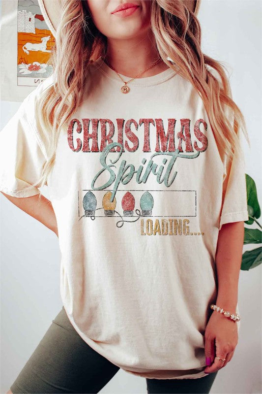 A person wearing a LOADING CHRISTMAS SPIRIT GRAPHIC TEE / T-SHIRT in beige, featuring the text "Christmas Spirit Loading..." and a festive holiday lights graphic, standing casually with one hand on their hip.