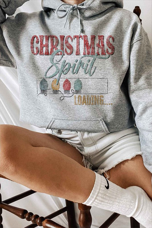 Person wearing a LOADING CHRISTMAS SPIRIT GRAPHIC HOODIE with "Christmas Spirit Loading..." text and three colorful Christmas lights, paired with denim shorts and white socks featuring a black swoosh logo.