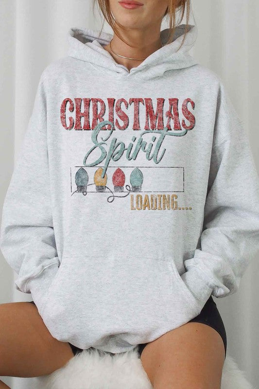 A person is wearing a gray LOADING CHRISTMAS SPIRIT HOODIE PLUS SIZE with "Christmas Spirit Loading..." and holiday lights printed on it, sitting with one knee up, and paired with white shorts and white socks. The hoodie features unisex sizing for a comfortable fit.