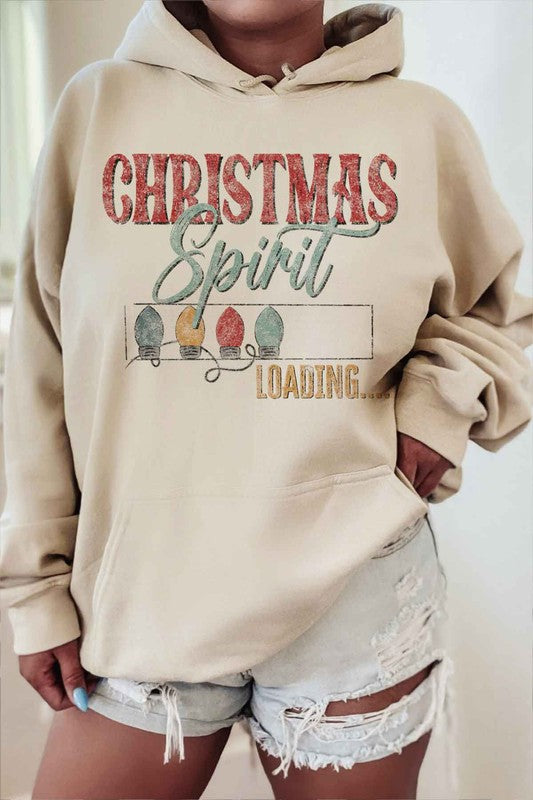 A person is wearing a gray LOADING CHRISTMAS SPIRIT HOODIE PLUS SIZE with "Christmas Spirit Loading..." and holiday lights printed on it, sitting with one knee up, and paired with white shorts and white socks. The hoodie features unisex sizing for a comfortable fit.
