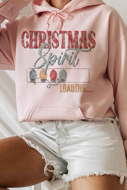 A person is wearing a gray LOADING CHRISTMAS SPIRIT HOODIE PLUS SIZE with "Christmas Spirit Loading..." and holiday lights printed on it, sitting with one knee up, and paired with white shorts and white socks. The hoodie features unisex sizing for a comfortable fit.