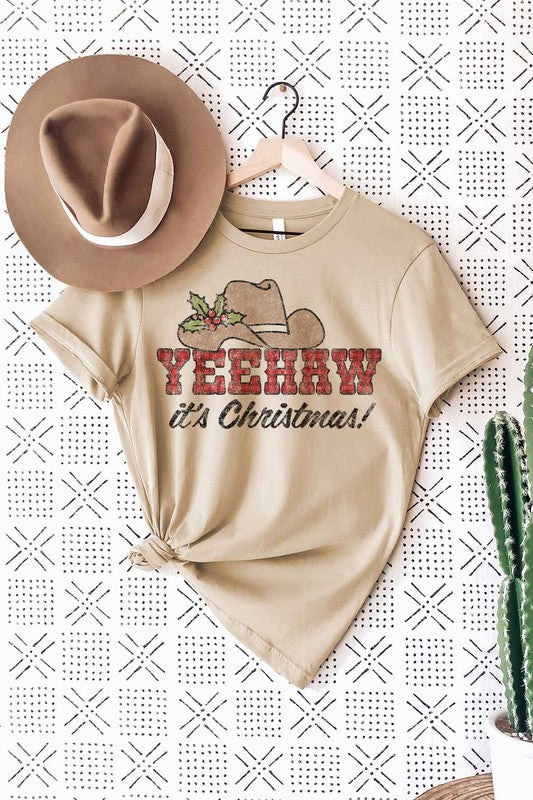 A woman wearing a pink hat and a white "YEEHAW COUNTRY CHRISTMAS GRAPHIC TEE / T-SHIRT" featuring a cowboy hat and holly design stands against a plain background, perfectly capturing the spirit of a YEEHAW Country Christmas. The unisex sizing guarantees comfort for anyone embracing the festive cheer.