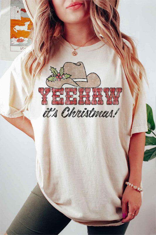 A beige t-shirt with the words "YEEHAW it's Christmas!" featuring a cowboy hat and holly decoration, hung on a hanger with a brown cowboy hat to the left and a cactus to the right. This YEEHAW COUNTRY CHRISTMAS GRAPHIC PLUS SIZE TEE is available in unisex sizing.