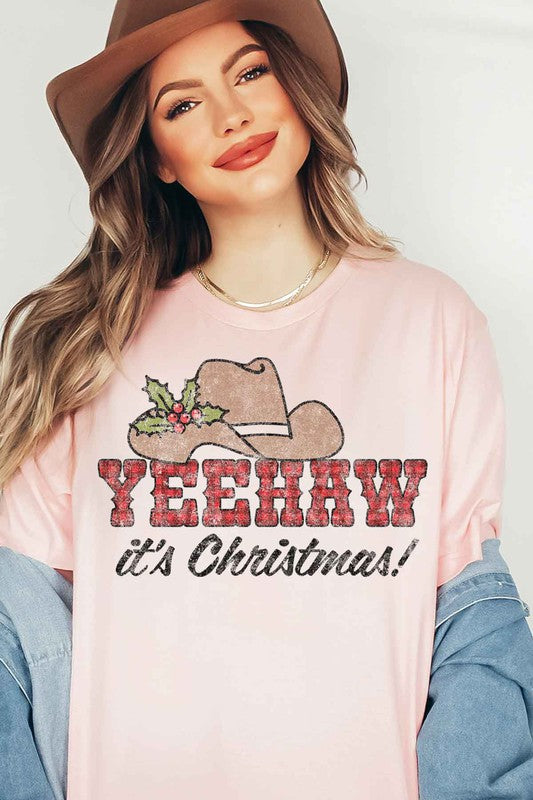 A beige t-shirt with the words "YEEHAW it's Christmas!" featuring a cowboy hat and holly decoration, hung on a hanger with a brown cowboy hat to the left and a cactus to the right. This YEEHAW COUNTRY CHRISTMAS GRAPHIC PLUS SIZE TEE is available in unisex sizing.