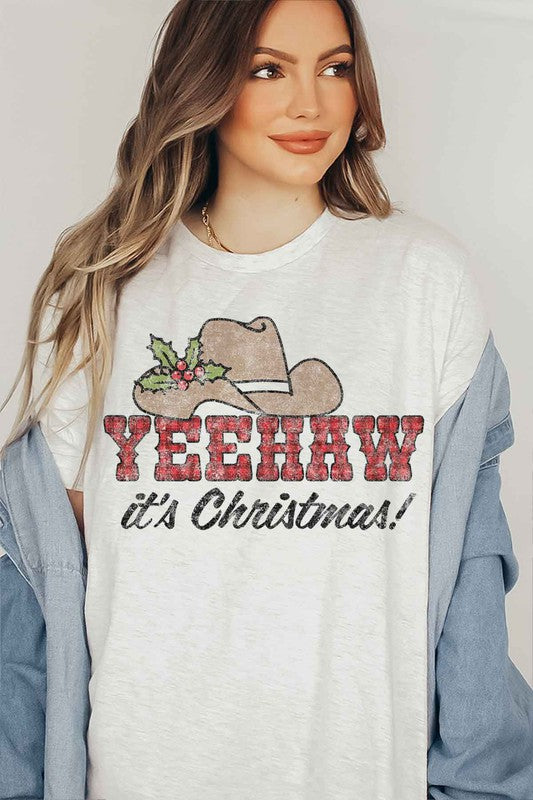 A beige t-shirt with the words "YEEHAW it's Christmas!" featuring a cowboy hat and holly decoration, hung on a hanger with a brown cowboy hat to the left and a cactus to the right. This YEEHAW COUNTRY CHRISTMAS GRAPHIC PLUS SIZE TEE is available in unisex sizing.