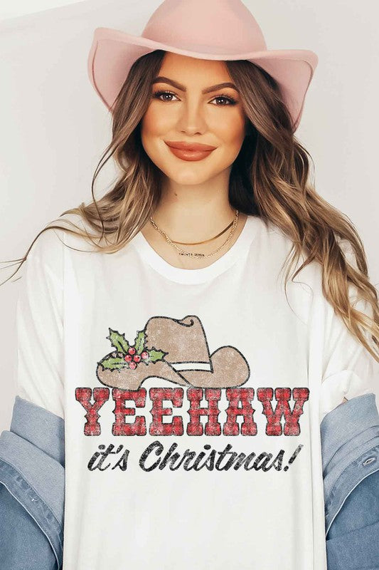 A beige t-shirt with the words "YEEHAW it's Christmas!" featuring a cowboy hat and holly decoration, hung on a hanger with a brown cowboy hat to the left and a cactus to the right. This YEEHAW COUNTRY CHRISTMAS GRAPHIC PLUS SIZE TEE is available in unisex sizing.