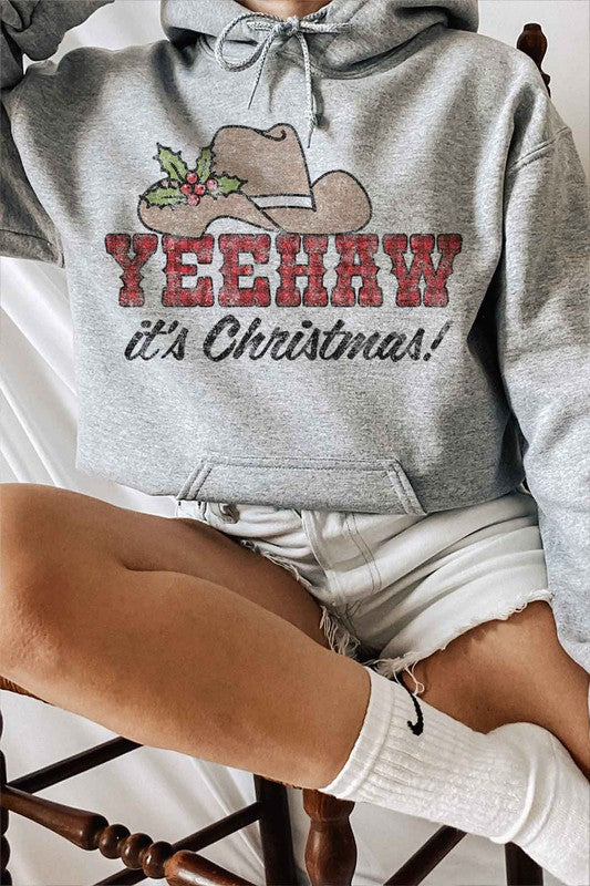 A person wearing the "YEEHAW COUNTRY CHRISTMAS GRAPHIC HOODIE" in beige, featuring a cowboy hat and holly graphic with the phrase "YEEHAW COUNTRY CHRISTMAS!" complemented by ripped denim shorts.