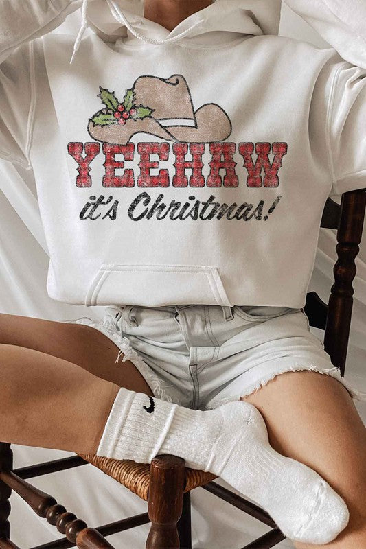 A person wearing the "YEEHAW COUNTRY CHRISTMAS GRAPHIC HOODIE" in beige, featuring a cowboy hat and holly graphic with the phrase "YEEHAW COUNTRY CHRISTMAS!" complemented by ripped denim shorts.