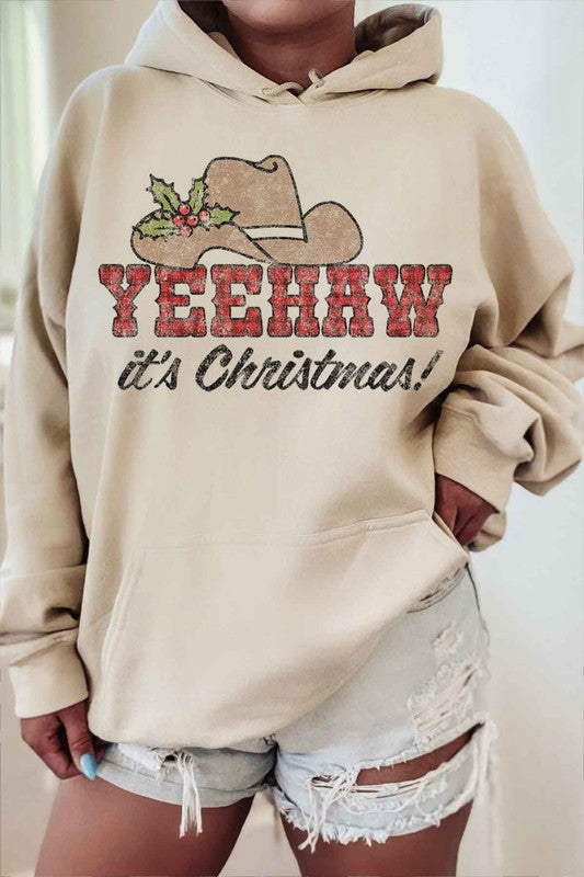 A person wearing the "YEEHAW COUNTRY CHRISTMAS GRAPHIC HOODIE" in beige, featuring a cowboy hat and holly graphic with the phrase "YEEHAW COUNTRY CHRISTMAS!" complemented by ripped denim shorts.