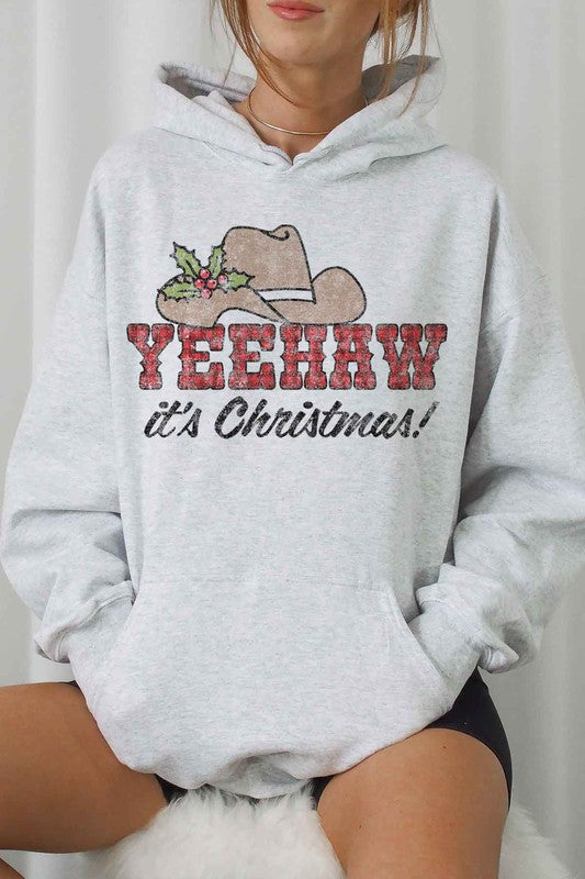 Person sitting on a wooden chair wearing a YEEHAW COUNTRY CHRISTMAS HOODIE PLUS SIZE, featuring a cowboy hat and holly graphic. They’re also sporting white socks with a Nike logo.