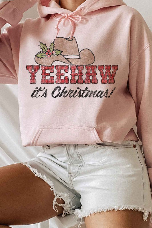 Person sitting on a wooden chair wearing a YEEHAW COUNTRY CHRISTMAS HOODIE PLUS SIZE, featuring a cowboy hat and holly graphic. They’re also sporting white socks with a Nike logo.