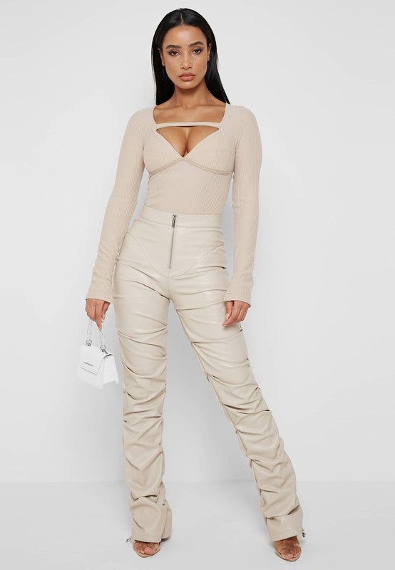 A woman stands against a plain white background, wearing a beige long-sleeve top with a cutout detail and high-waisted SEXY PU LEATHER PANTS in beige. She holds a small white handbag, embracing the skinny style, and has long dark hair.