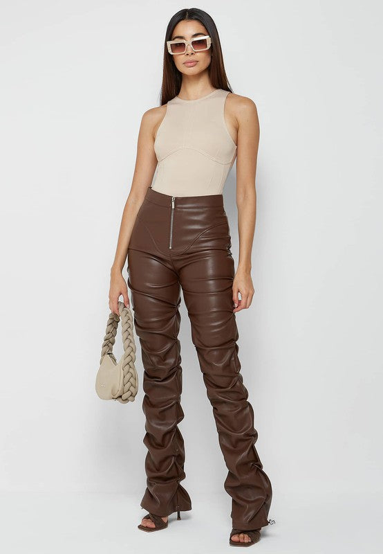 A person stands against a plain background wearing a sleeveless beige top, the SEXY PU LEATHER PANTS in high-waisted brown skinny style, white-framed sunglasses, and holding a beige braided handbag.