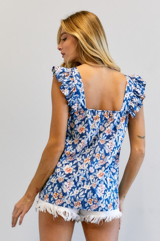 A woman in a vibrant summer wardrobe stands against a plain background, wearing the Floral Printed Ruffle Sleeveless Top paired with white shorts, with one hand casually tucked in her pocket.